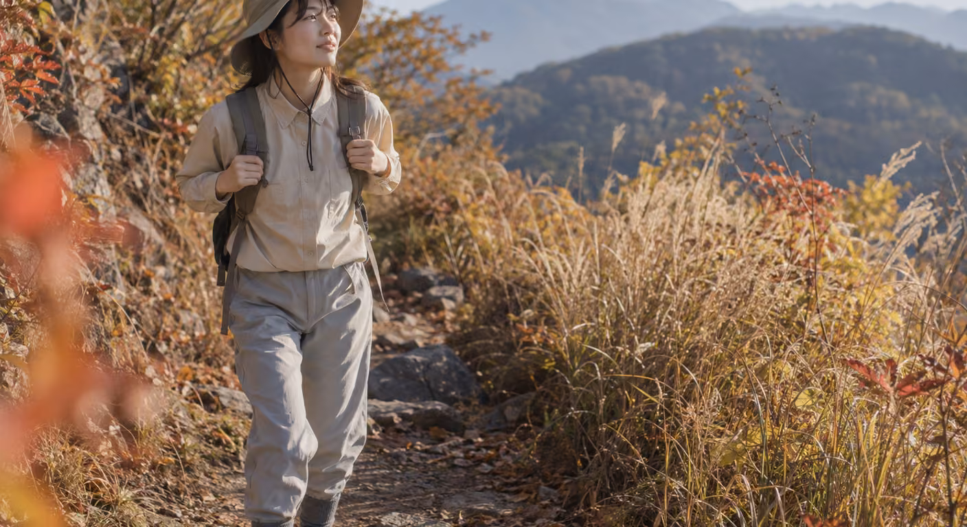 秋の山道の入り口で、袖口を留めた明るい色の長袖シャツと、裾を靴下に入れた薄いグレーのパンツの女性が歩き出すところ