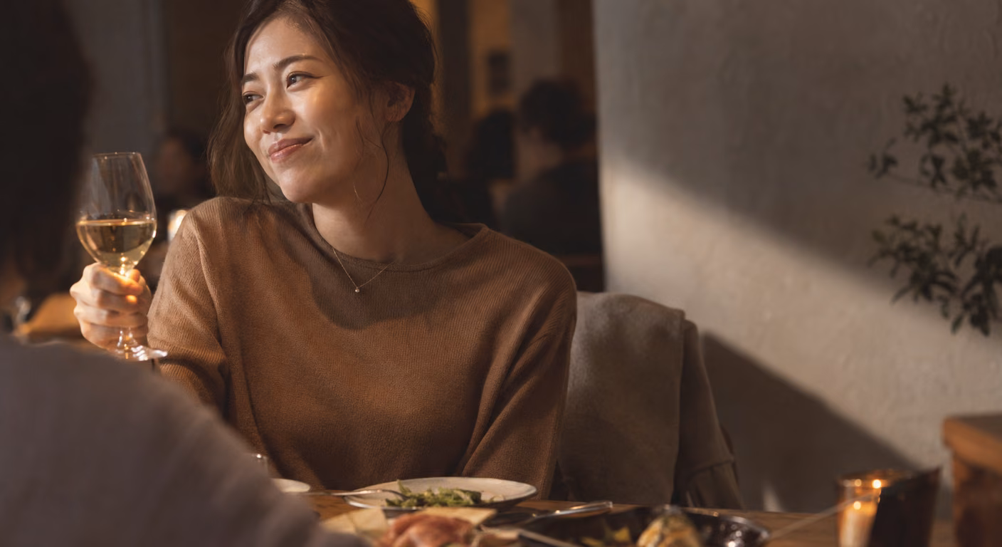 暖かい照明のレストランで、キャメルの細い編みのニットに細い首飾りの女性が、向かいの人にグラスを掲げている