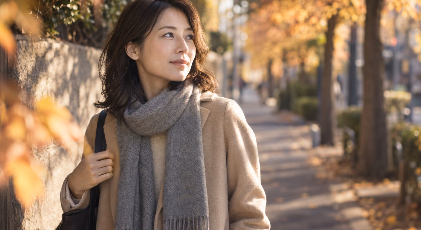 グレーの細いマフラーを首にひと巻きして両端を胸の前に縦に垂らした女性が、秋の並木道で横を向いて立ち止まっている