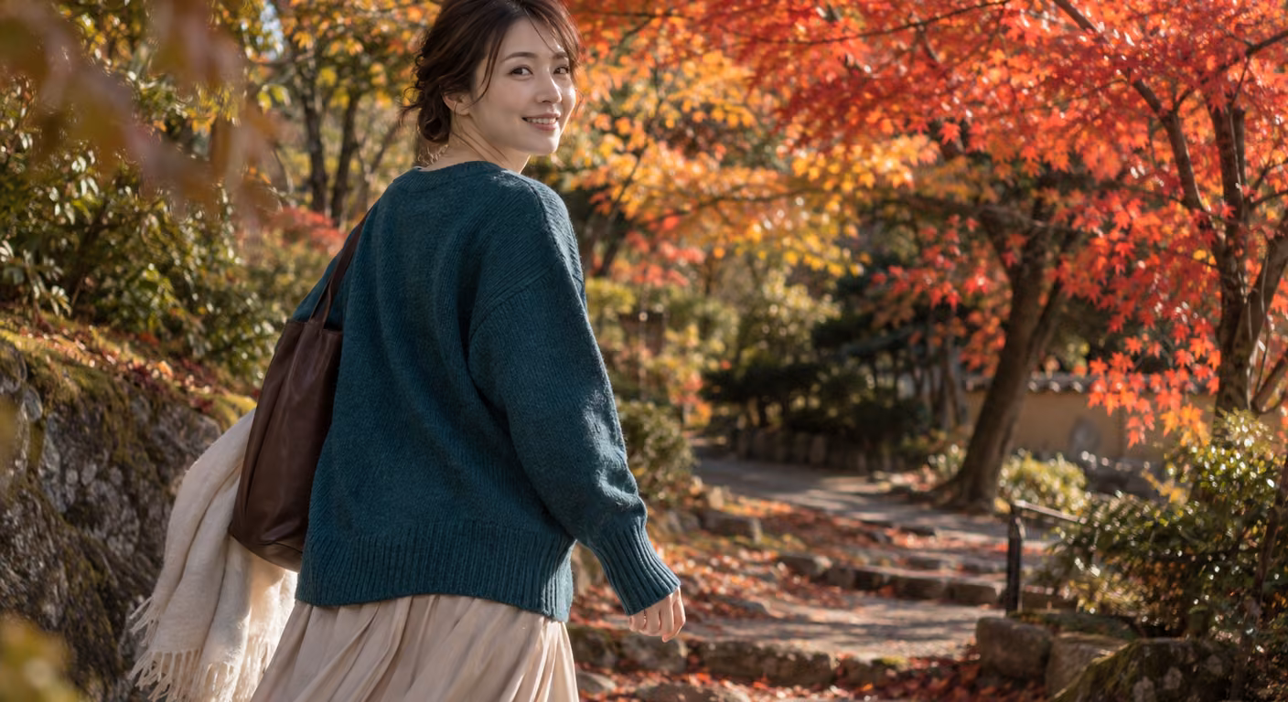赤く色づいたもみじの下で、深い青緑のニットを着た女性が振り返りながら歩いている秋の庭の小道