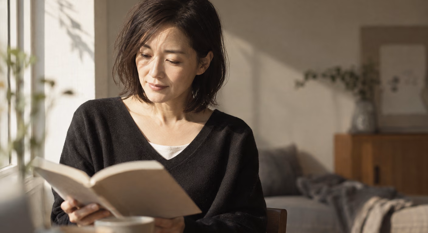 黒いVネックのニットの襟元から生成りのインナーを少しのぞかせた女性が、窓の光を横から受けながら手元の本に目を落としている