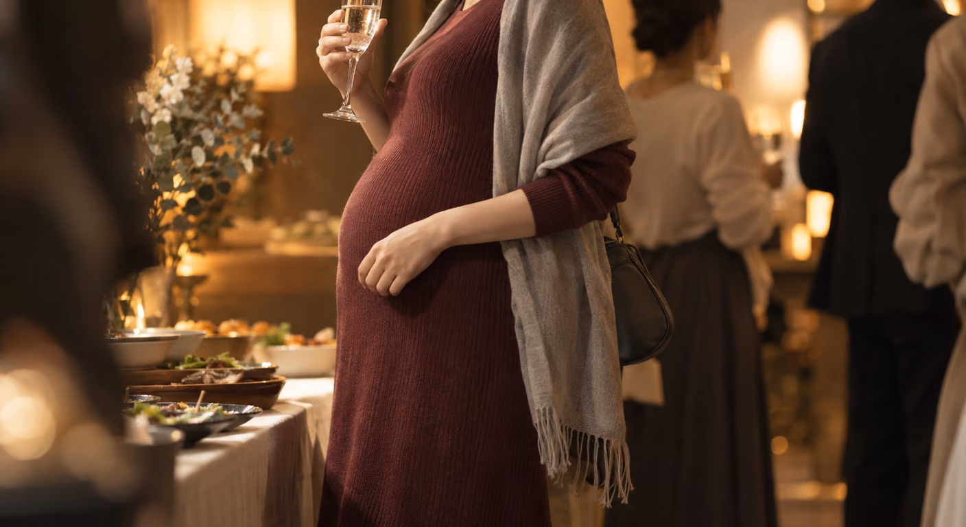 夜のレストランの立食パーティで、グラスを片手に横を向いて立つ妊婦の首元から膝。体に沿う深いボルドーの長袖ニットドレスがお腹の丸みに沿って落ち、肩にグレーのショールを掛けて、もう一方の手をお腹に添えている。手前には料理の並ぶテーブル、奥にはぼけた照明と人の影