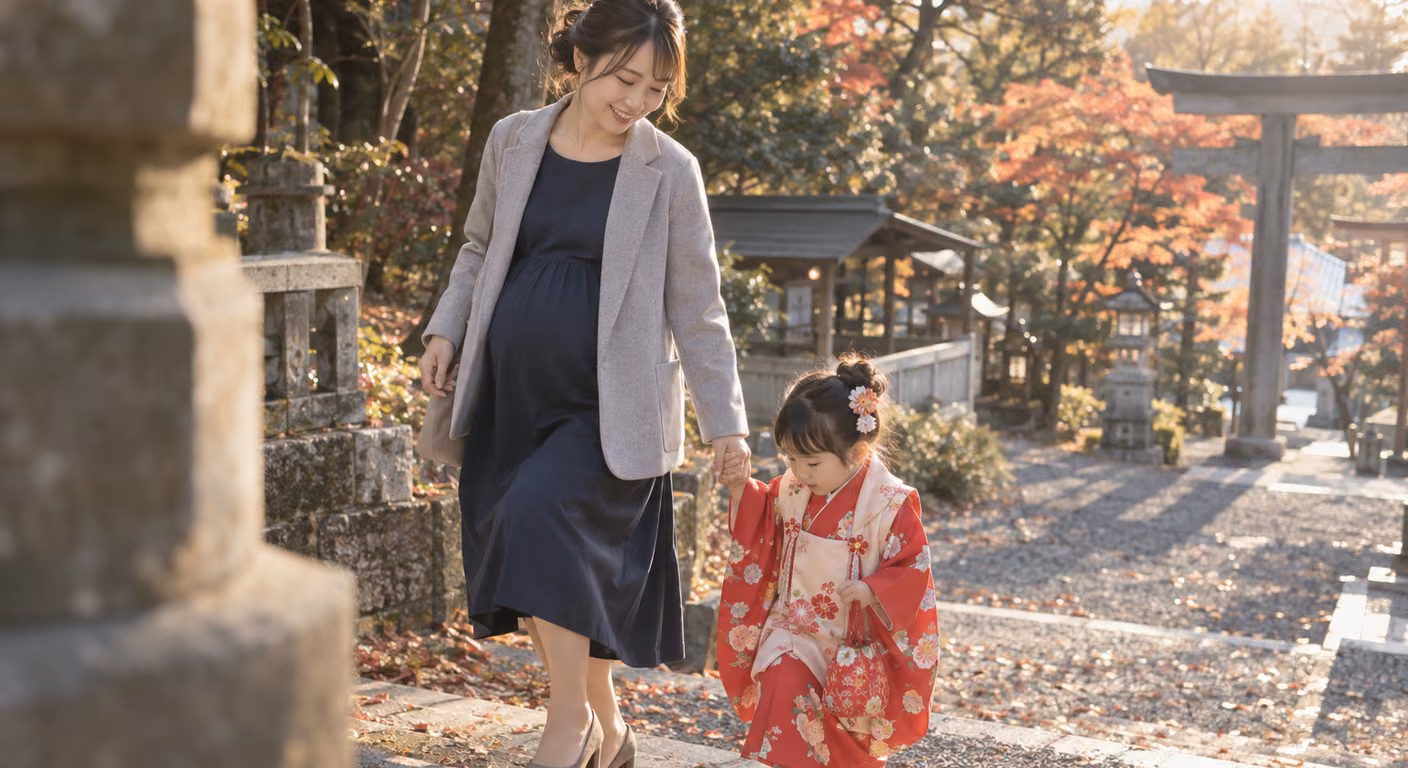 秋の神社の参道で、お腹のふくらんだ女性が、紺のワンピースに前を開けた灰色の羽織りを重ね、赤い着物の子どもの手を引いて歩いている