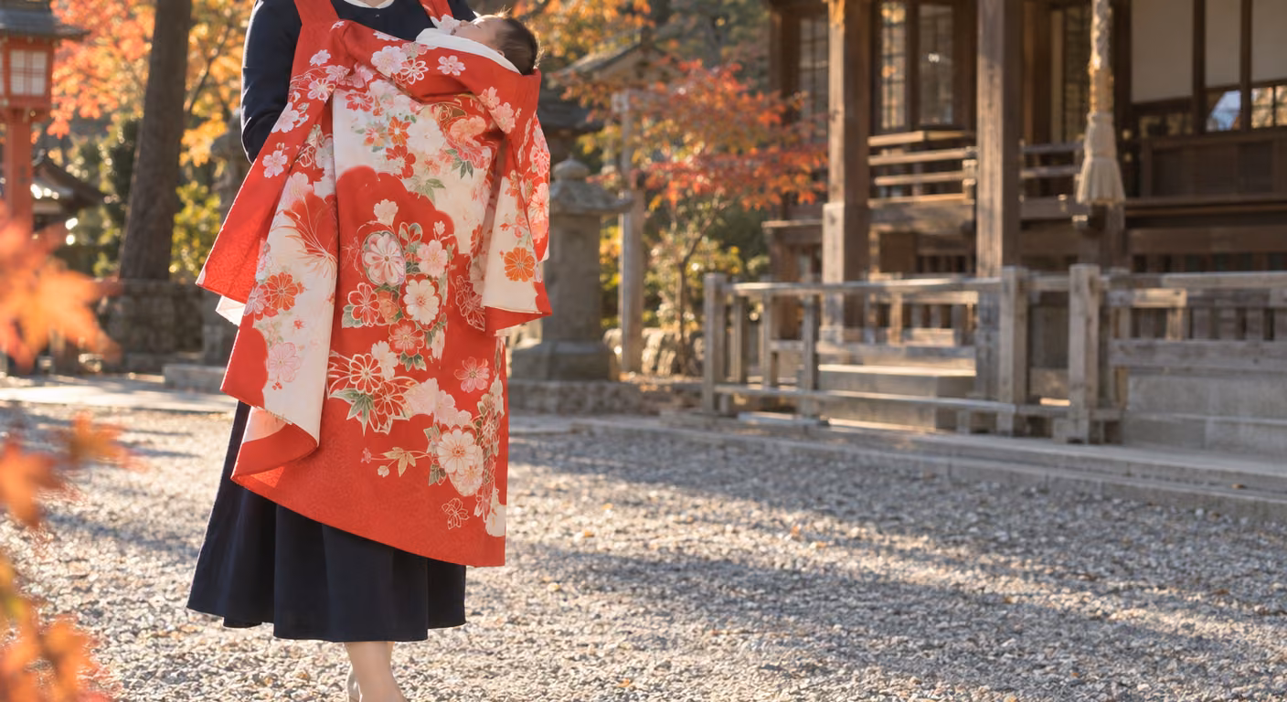 紅葉した神社の砂利の参道で、赤い花柄の祝い着を掛けた赤ちゃんを抱く女性。胸から膝のあたりまでが祝い着で覆われ、その下から紺のワンピースの裾が出ている