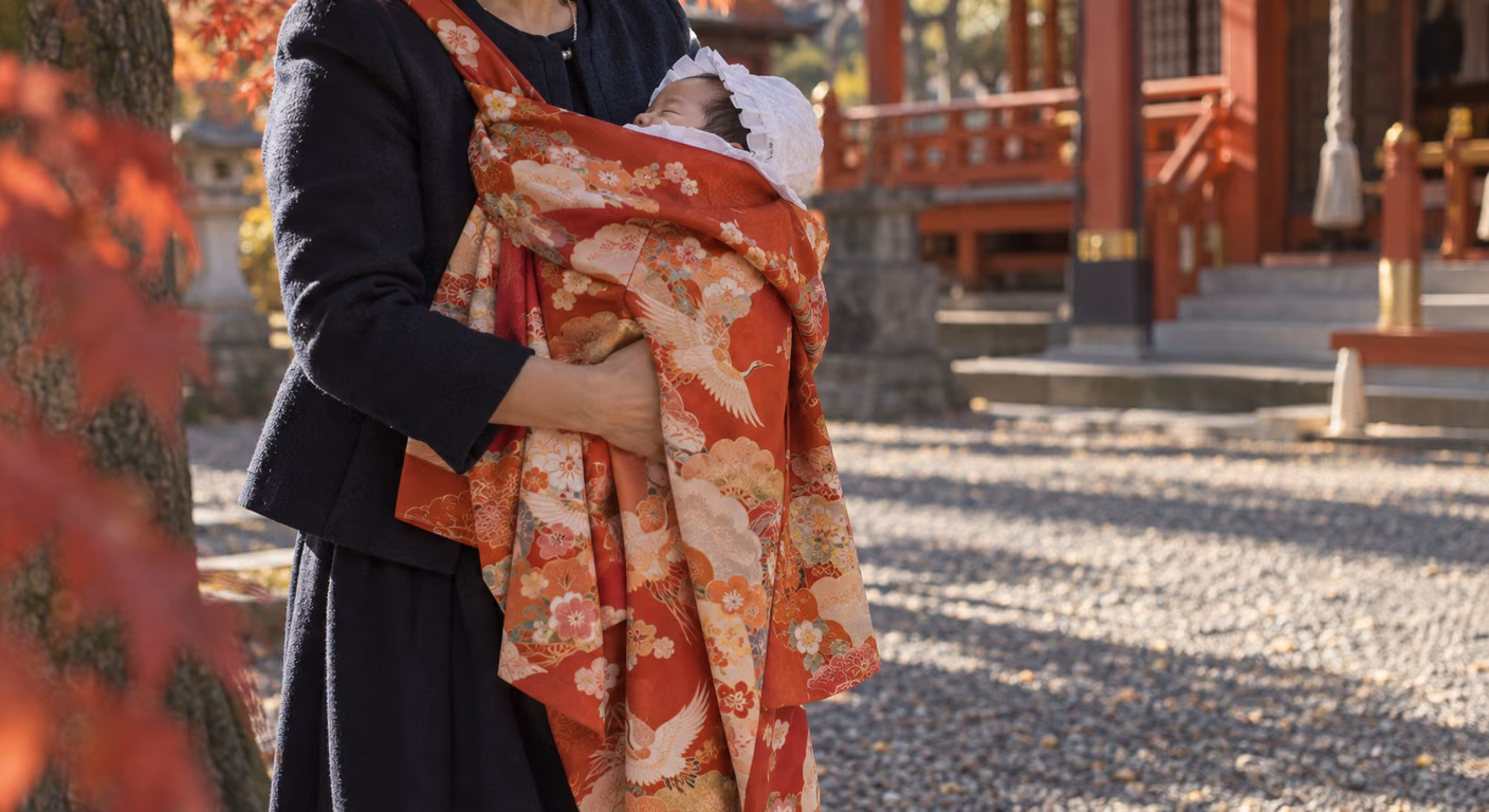 紅葉の神社の境内で、赤い祝い着を掛けた赤ちゃんを抱く女性の胸元から膝。祝い着の端が濃紺のジャケットの肩にかかっている